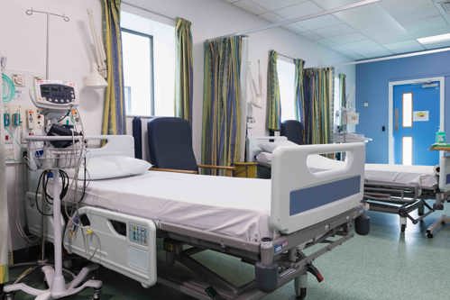 A photo of the Clinical research Facility. In the photo A bright hospital ward with two empty patient beds separated by patterned privacy curtains. Medical monitoring equipment is positioned beside one bed, while blue clinical chairs sit next to the beds.